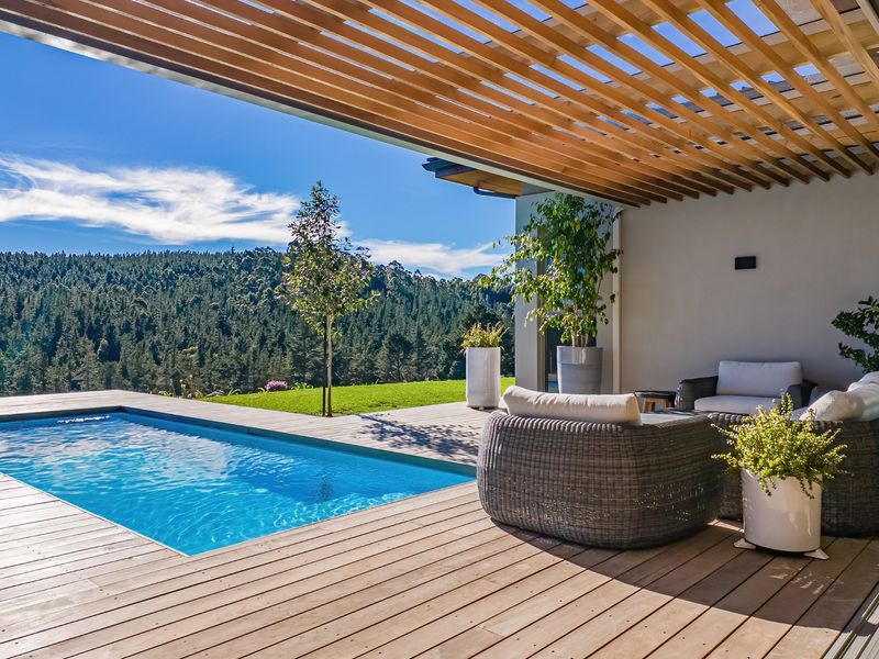 A beautifully maintained outdoor pool area featuring timber decking, a modern wooden pergola, and scenic views of the surrounding forest.