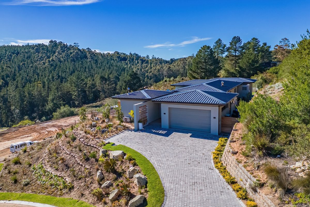 Aerial view of a modern house featuring a metal roof, paved driveway, double garage, and landscaped terraced gardens set against a forested hillside.