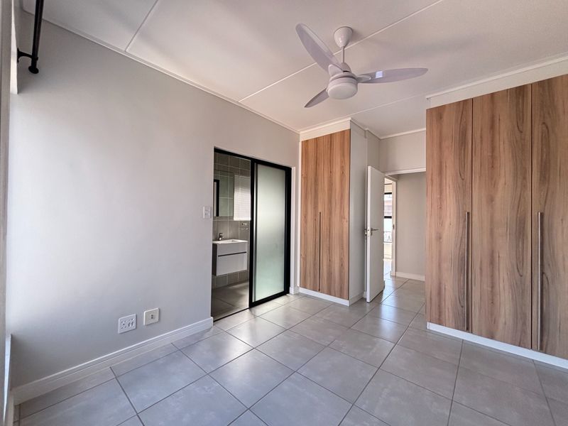 A bright, modern bedroom featuring neutral tiled flooring, built-in wooden cupboards, a ceiling fan, and an en-suite bathroom access.