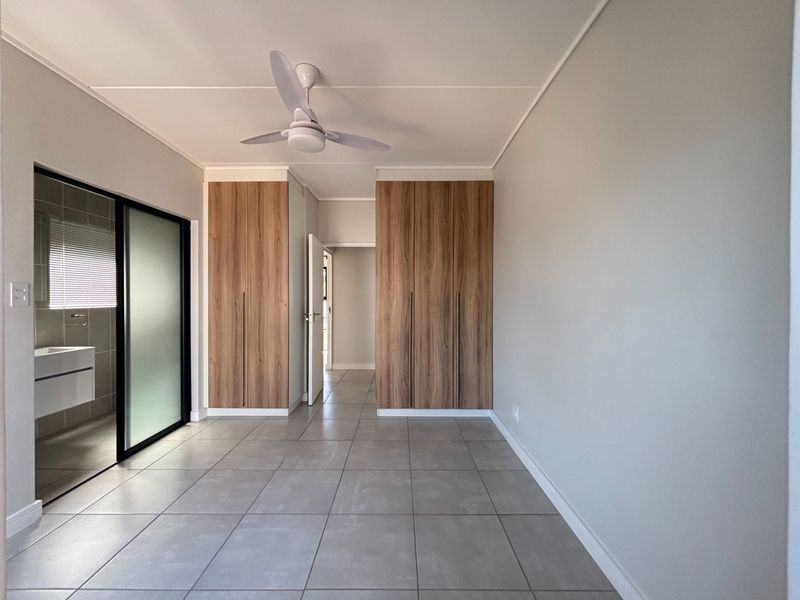 A modern, empty bedroom featuring neutral tiled flooring, built-in wood-finish cupboards, a ceiling fan, and direct access to an en-suite bathroom.