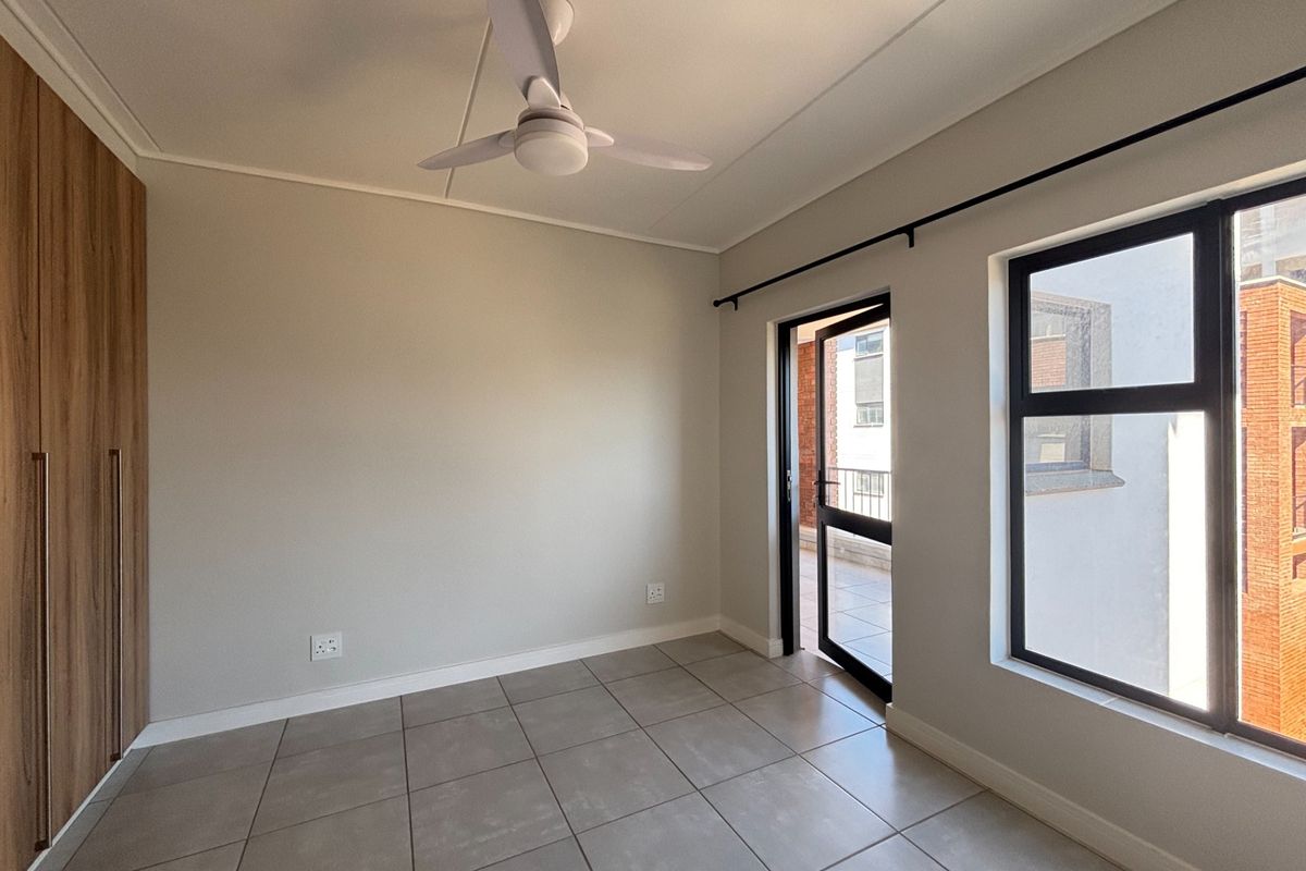 A bright, empty bedroom featuring modern tiled flooring, built-in wooden cupboards, a ceiling fan, and direct access to an outdoor balcony area.
