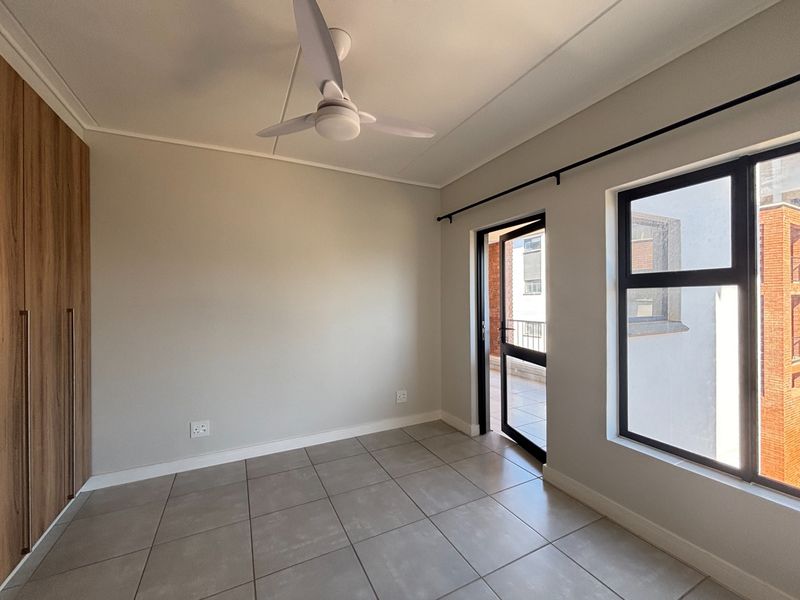 A bright, empty bedroom featuring modern tiled flooring, built-in wooden cupboards, a ceiling fan, and direct access to an outdoor balcony area.