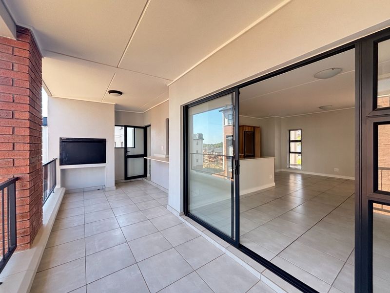 Spacious covered balcony featuring a built-in braai, tiled flooring, and large sliding doors providing seamless access to the open-plan interior living area.