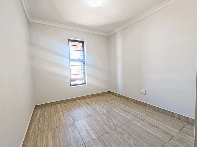 A clean, empty bedroom featuring neutral tiled flooring, coving, and a window fitted with security bars.
