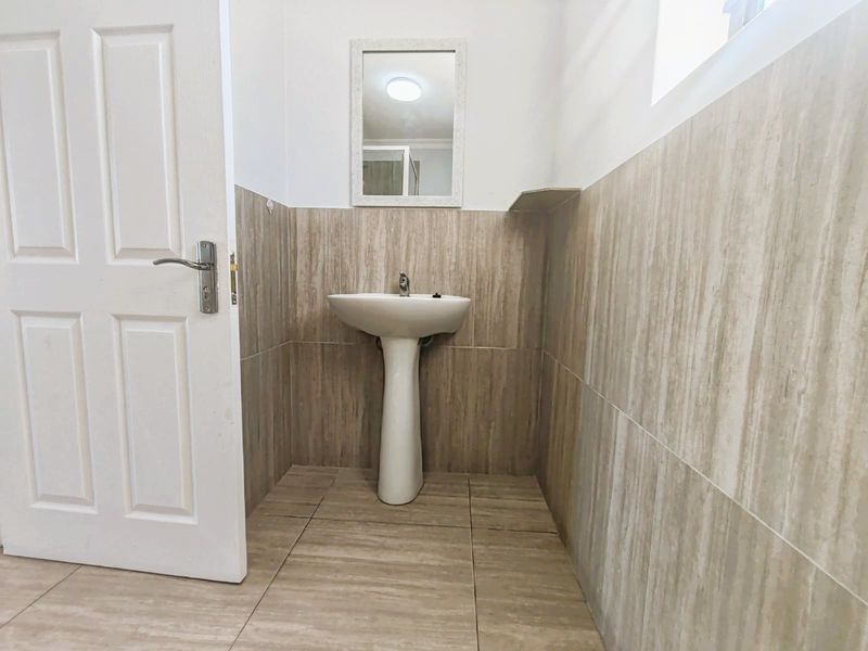 A clean, minimalist bathroom featuring a pedestal basin, neutral-toned wall and floor tiling, and a wall-mounted mirror.