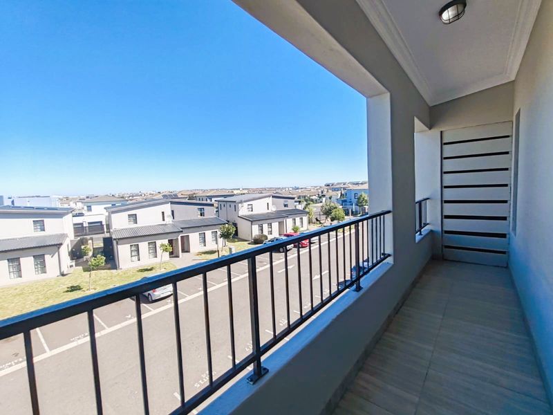 A clean, covered balcony featuring tiled flooring, a metal balustrade, and a privacy screen, offering an elevated view of the surrounding residential complex.