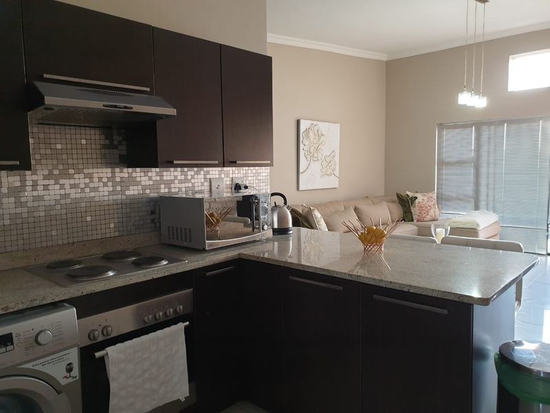 Open-plan kitchen and living area featuring dark cabinetry, granite countertops, mosaic backsplash, and integrated appliances, flowing into a bright, neutrally decorated living space.
