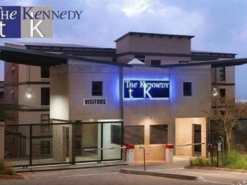 The main entrance and security gatehouse of The Kennedy apartment complex, featuring modern signage, controlled access boom gates, and exterior lighting.