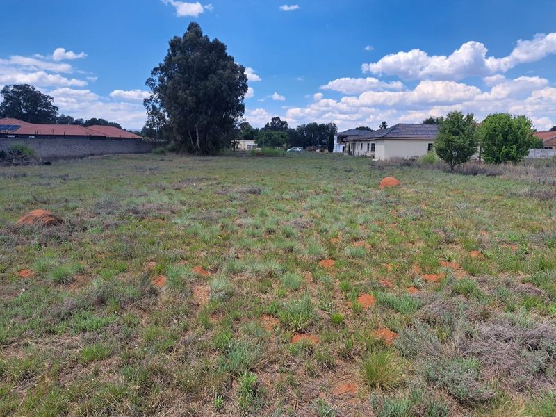 A vacant residential plot featuring natural grass cover, perimeter boundary walls, and mature trees, situated in an established residential neighborhood.