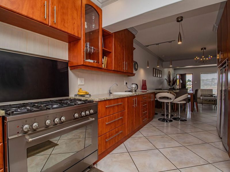 Open-plan kitchen featuring warm-toned wooden cabinetry, granite countertops, and a professional-style gas stove, flowing seamlessly into the adjacent dining and living areas.
