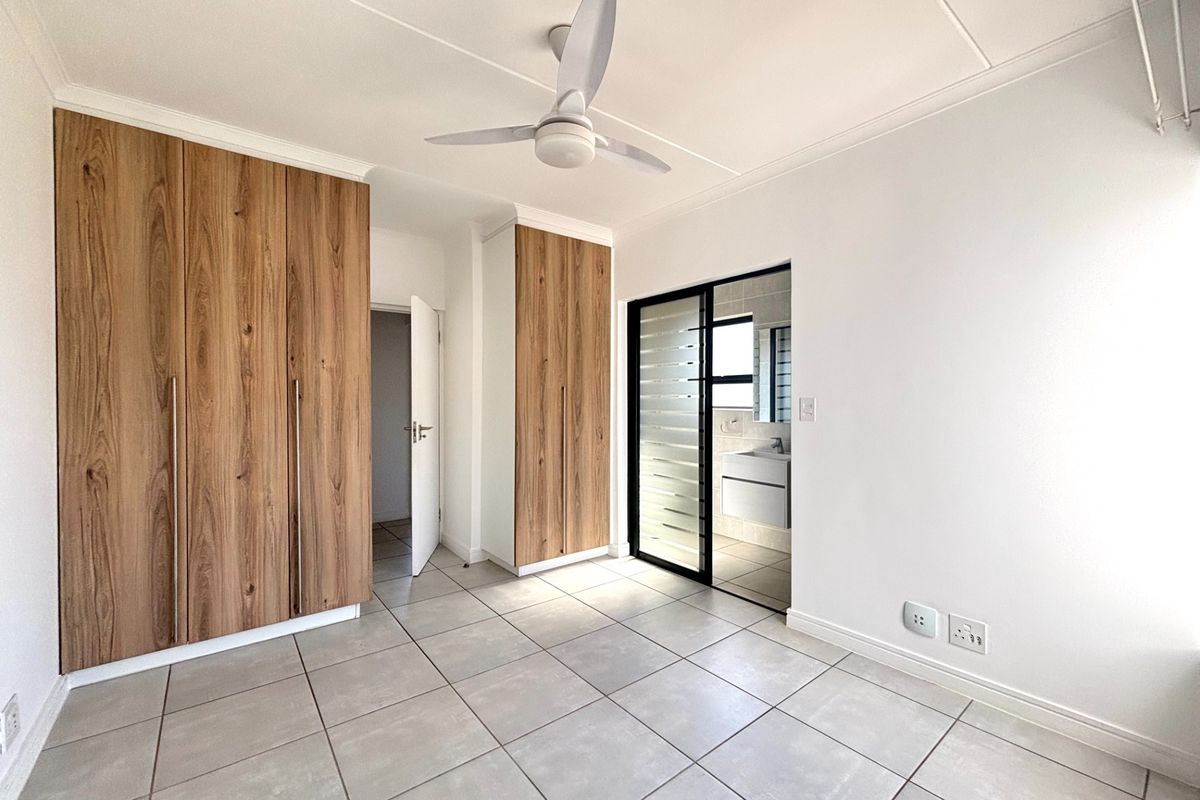 A bright, modern bedroom featuring neutral tiled flooring, ample built-in wooden cupboards, a ceiling fan, and direct access to an en-suite bathroom.