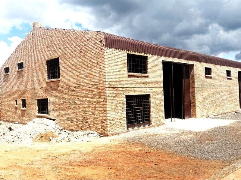 Industrial warehouse building featuring face brick construction, a curved metal roof, and secure window grilles, set on an unfinished site.