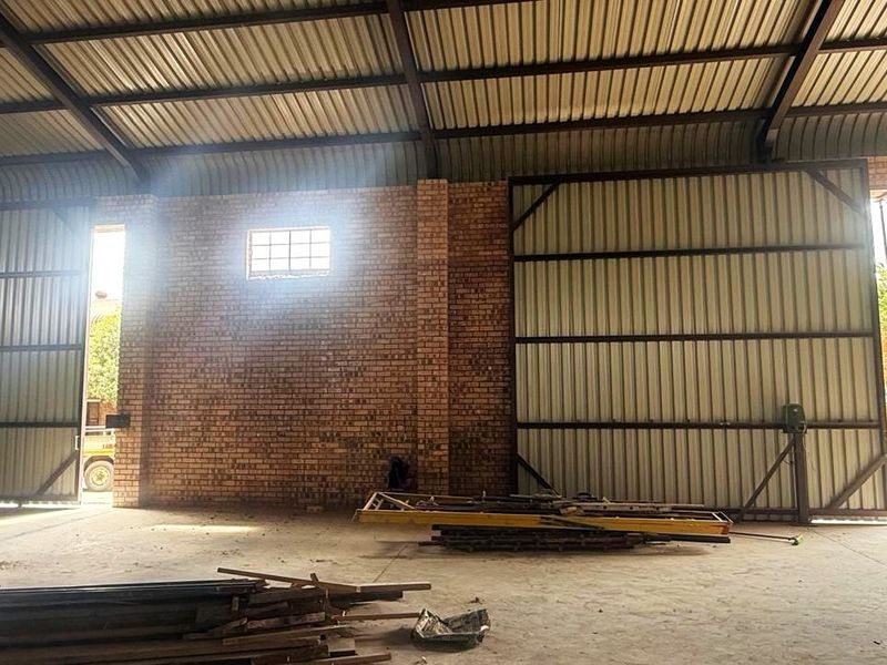 Spacious industrial warehouse or garage featuring exposed brick walls, high corrugated metal roofing with steel trusses, and large sliding access doors.
