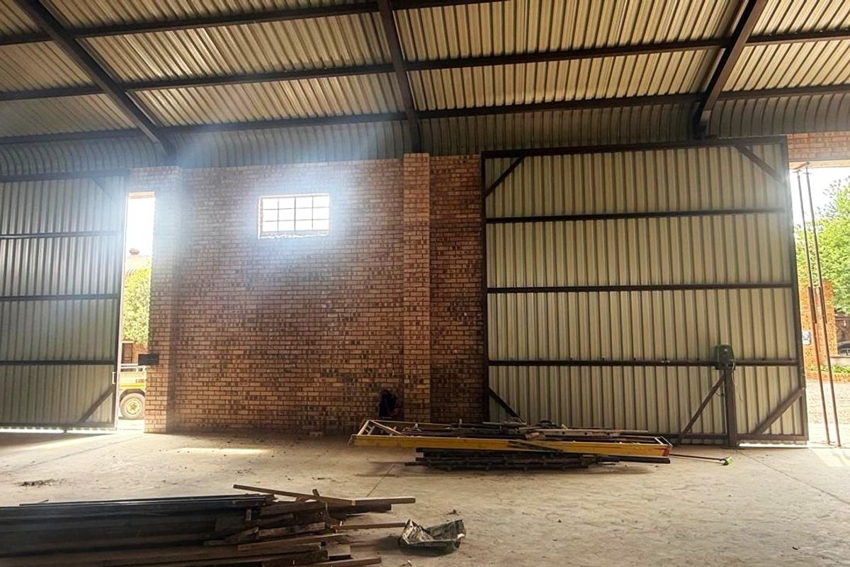 Spacious industrial warehouse or garage featuring exposed brick walls, high corrugated metal roofing with steel trusses, and large sliding access doors.