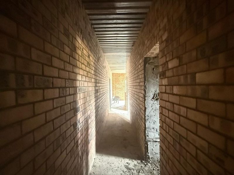 A narrow industrial-style hallway under construction featuring exposed brick walls and a corrugated metal ceiling, currently in a shell condition.