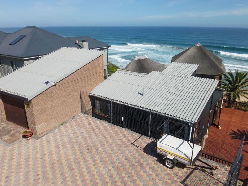 Aerial view of a coastal property featuring a brick-paved driveway, garage, corrugated metal roofing, and a timber deck overlooking the ocean.