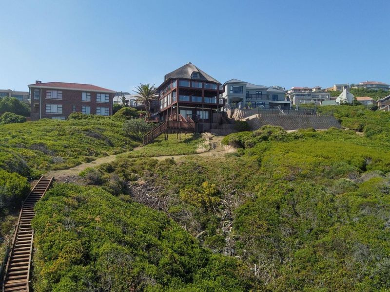 Aerial view of a coastal property featuring a multi-level home with a thatched roof, wooden decking, and a private staircase leading through the landscape.