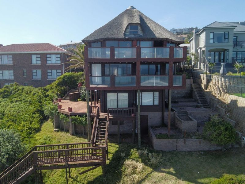 Multi-level rear elevation featuring a thatched roof, extensive timber decking, glass balustrades, and tiered retaining walls set within a coastal landscape.