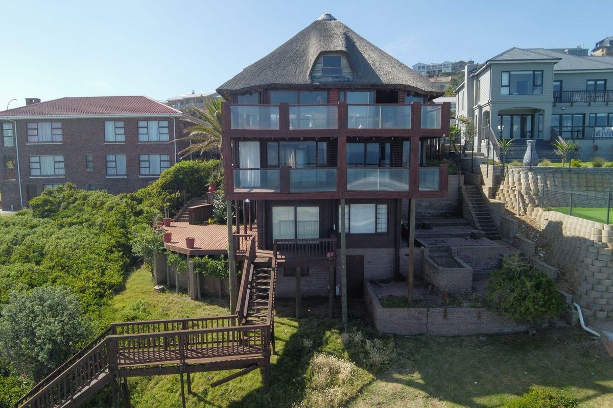 Multi-level rear elevation featuring a thatched roof, extensive timber decking, glass balustrades, and tiered retaining walls set within a coastal landscape.