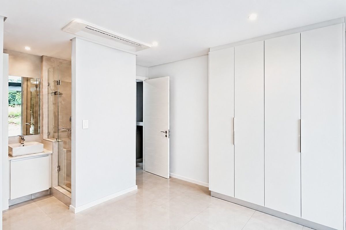 A modern, minimalist bedroom featuring built-in cupboards, tiled flooring, and an integrated en-suite bathroom with a glass shower enclosure.