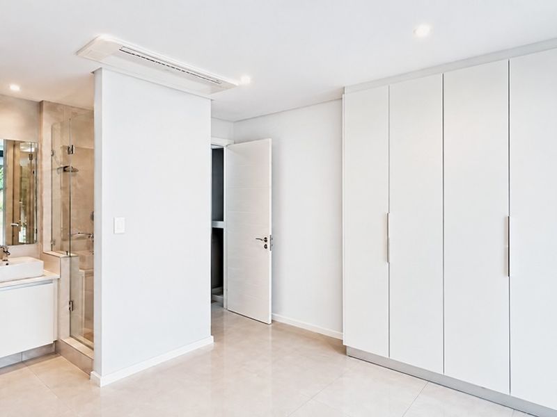 A modern, minimalist bedroom featuring built-in cupboards, tiled flooring, and an integrated en-suite bathroom with a glass shower enclosure.