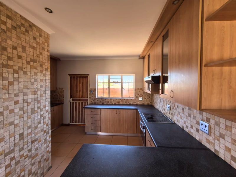 A functional kitchen featuring wood-finish cabinetry, dark laminate countertops, mosaic tile splashbacks, and integrated appliances, presented in a clean, empty space.