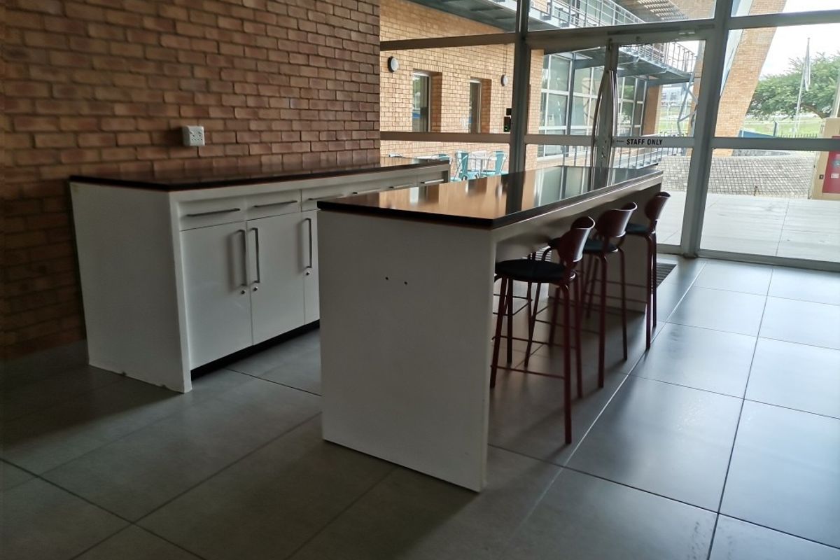 Modern commercial kitchenette area featuring built-in cabinetry, laminate countertops, and large format floor tiles, adjacent to a full-height glass entrance.