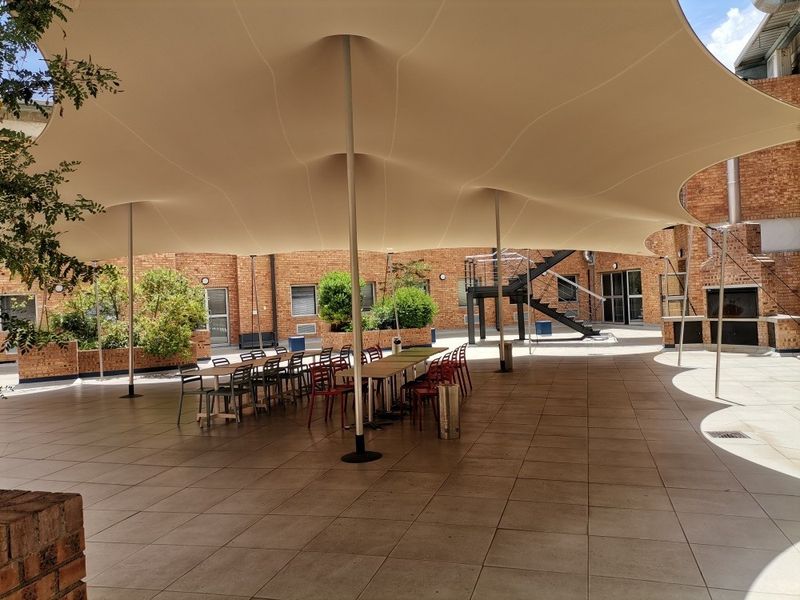 A spacious outdoor courtyard featuring a large tensile fabric canopy, tiled flooring, and exposed brick architecture, suitable for commercial or communal use.