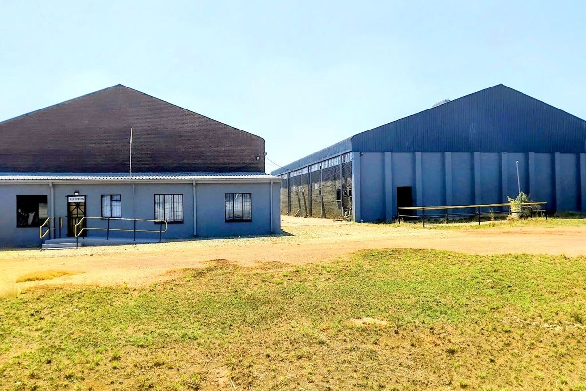 Exterior view of a commercial warehouse facility featuring a reception entrance, metal cladding, and a large unpaved yard area.