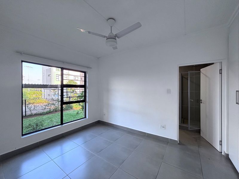 A bright, empty bedroom featuring neutral tiled flooring, a ceiling fan, and an en-suite bathroom, offering a clean canvas for personalization.