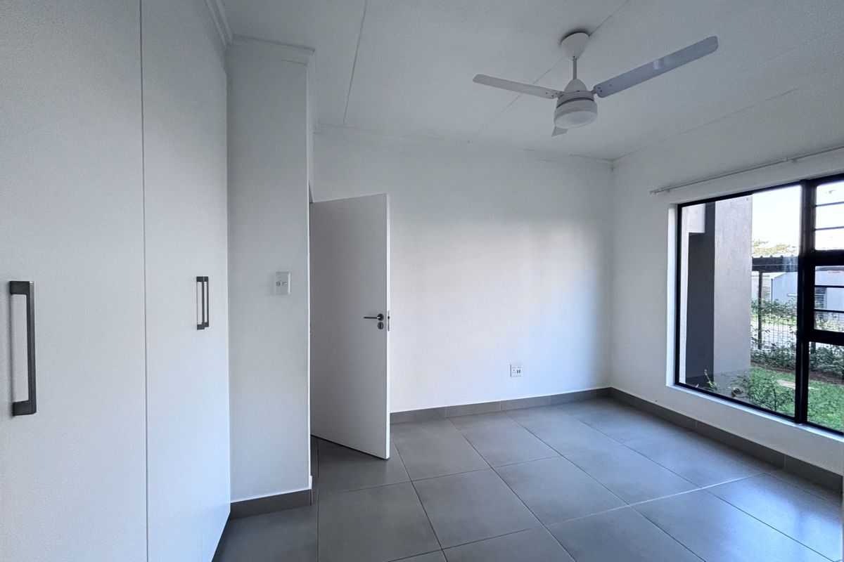A bright, empty bedroom featuring modern grey tiled flooring, built-in cupboards, and a ceiling fan, offering a clean and neutral living space.