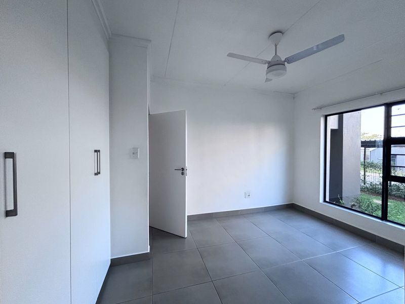 A bright, empty bedroom featuring modern grey tiled flooring, built-in cupboards, and a ceiling fan, offering a clean and neutral living space.
