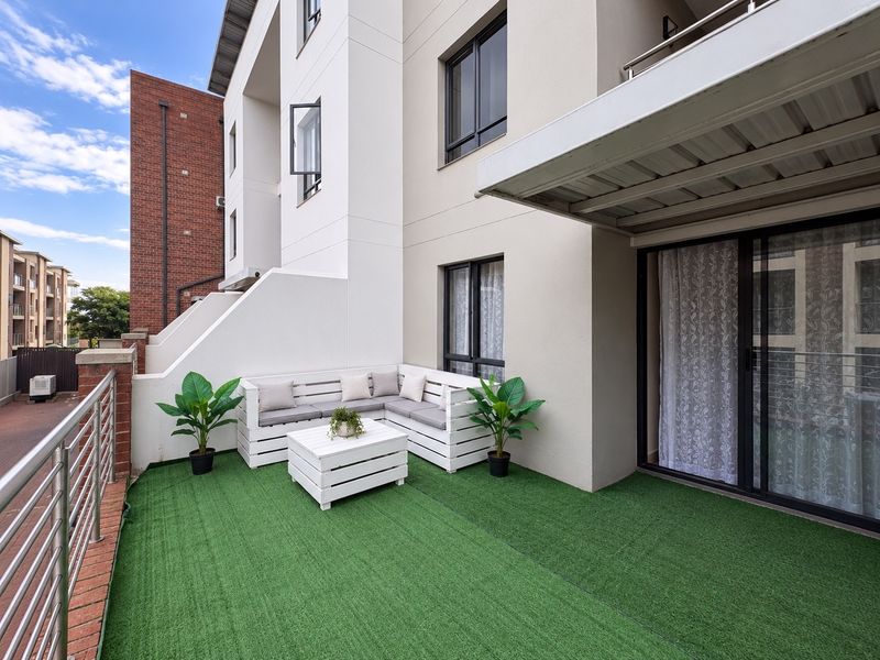 A well-maintained apartment balcony featuring artificial turf flooring, a covered overhead structure, and secure metal railings with access via sliding glass doors.