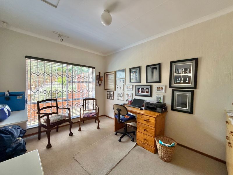 A functional home office space featuring a large window with security bars, neutral carpeting, and standard ceiling lighting.