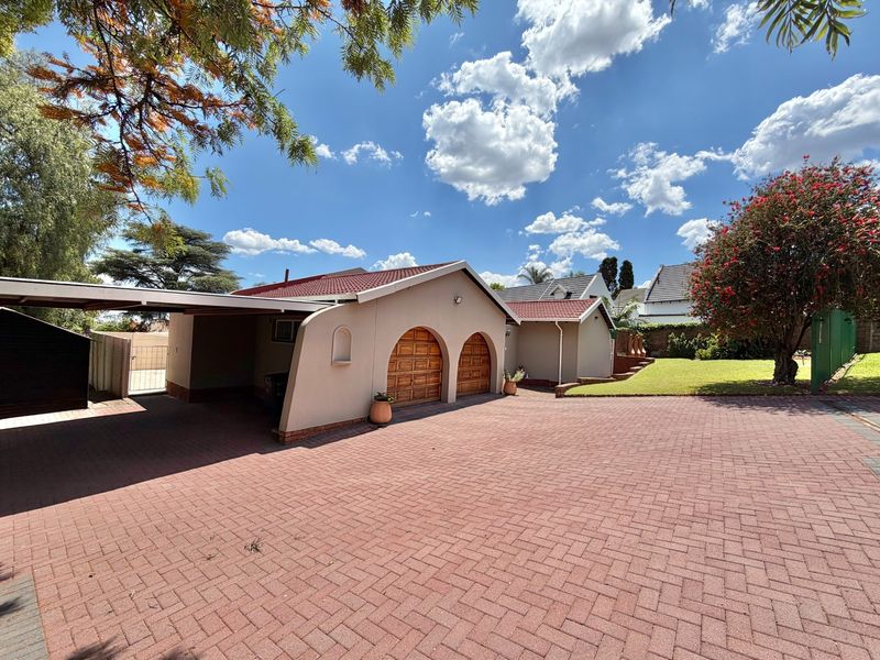 Front view of the property featuring a paved driveway, double garage with arched wooden doors, and an attached carport.