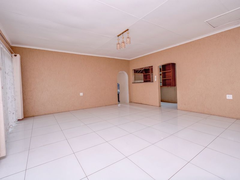 A spacious, empty living area featuring neutral textured walls, white tiled flooring, and an archway leading to a kitchen area with a serving hatch.