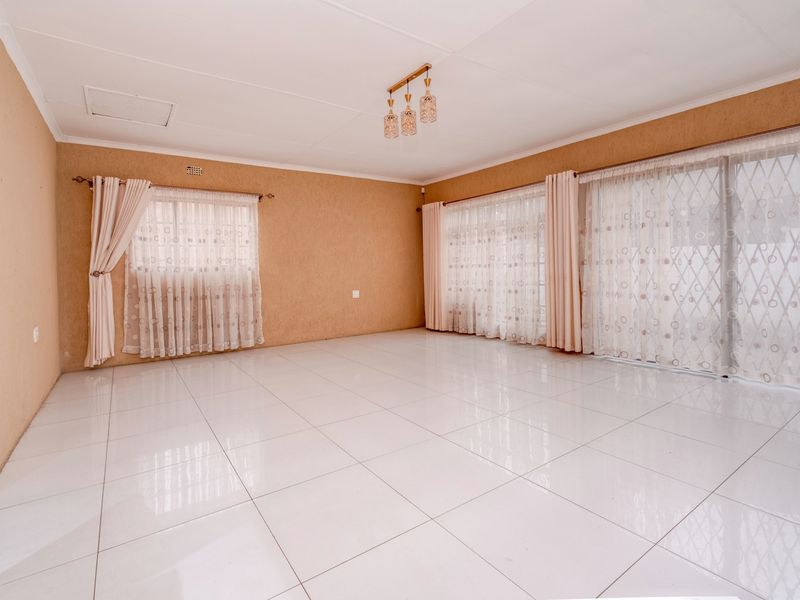 A spacious, empty living room featuring bright white tiled flooring, neutral textured walls, and large windows with sliding glass doors leading outside.