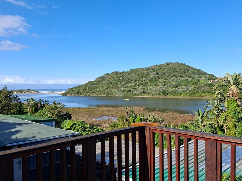 Scenic elevated view overlooking a tranquil lagoon, lush green hills, and distant coastline, framed by a wooden balcony railing.