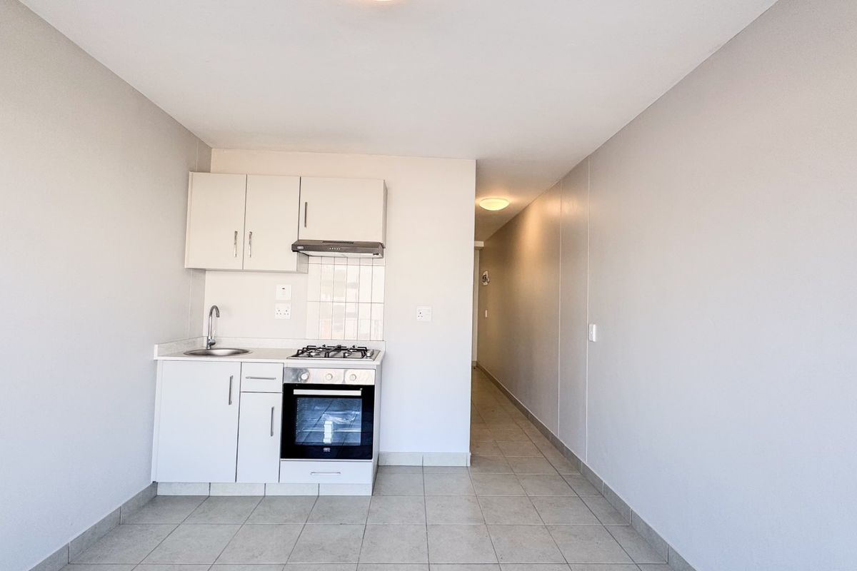 A compact, clean kitchen featuring white built-in cabinetry, a gas hob, oven, extractor fan, and tiled flooring, leading into a hallway.