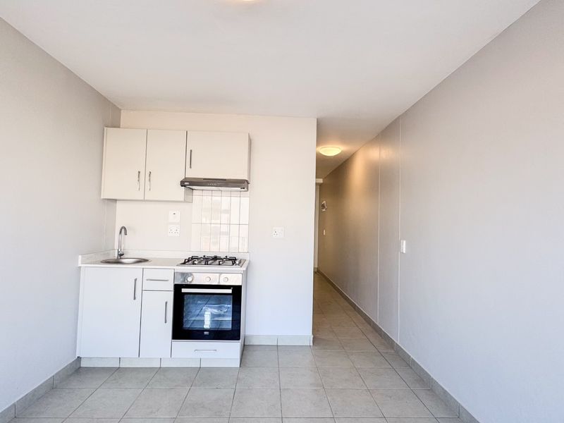 A compact, clean kitchen featuring white built-in cabinetry, a gas hob, oven, extractor fan, and tiled flooring, leading into a hallway.