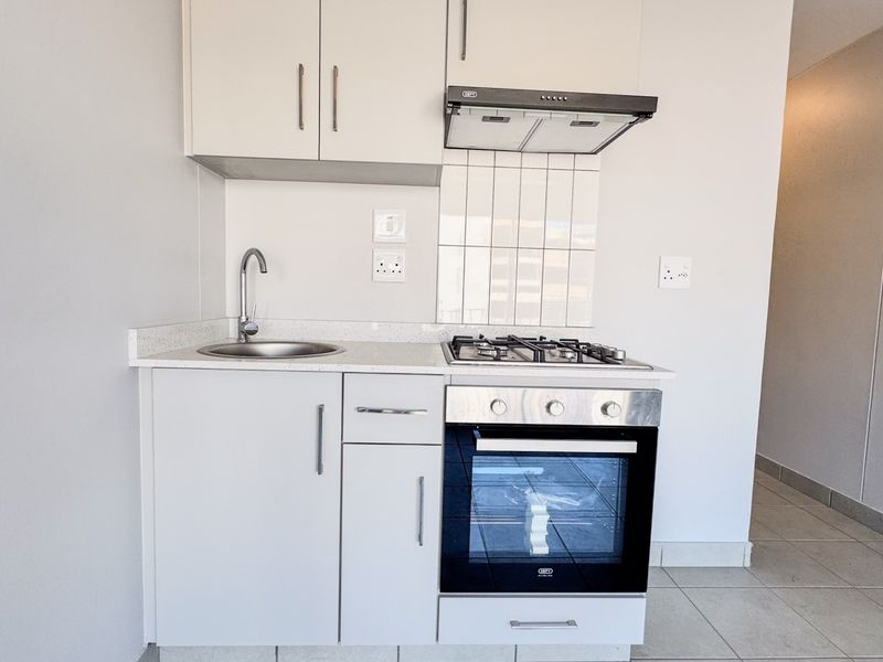A compact, modern kitchen featuring white cabinetry, a gas hob, built-in oven, extractor fan, and a stainless steel sink with a stone countertop.
