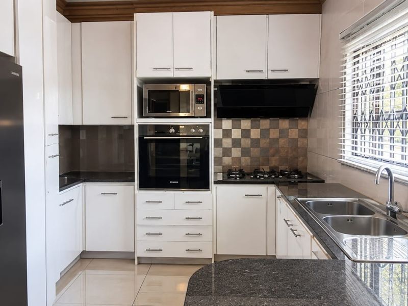 Modern kitchen featuring white cabinetry, granite countertops, built-in oven, microwave, gas hob, and a double sink with tiled backsplash and flooring.