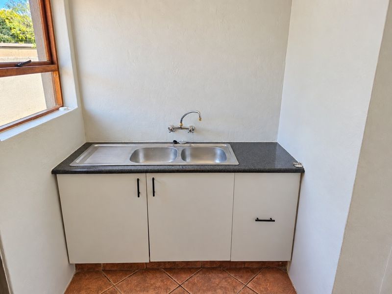 A clean and functional scullery area featuring a stainless steel double sink, granite countertop, and built-in cabinetry with tiled flooring.
