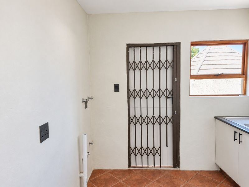 A functional laundry area featuring terracotta floor tiles, plumbing connections for appliances, and a secure metal gate leading to the exterior.