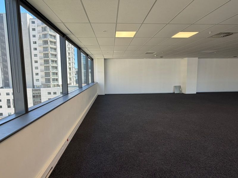A spacious, empty open-plan office area featuring large windows with city views, a suspended ceiling with recessed lighting, and dark carpet flooring.