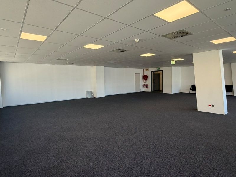 A spacious, open-plan office area featuring a suspended ceiling with recessed lighting, air conditioning vents, and dark carpeting throughout.
