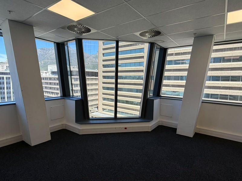A bright, empty corner office space featuring large windows with city views, suspended ceilings, and dark carpeting, suitable for a professional workspace.