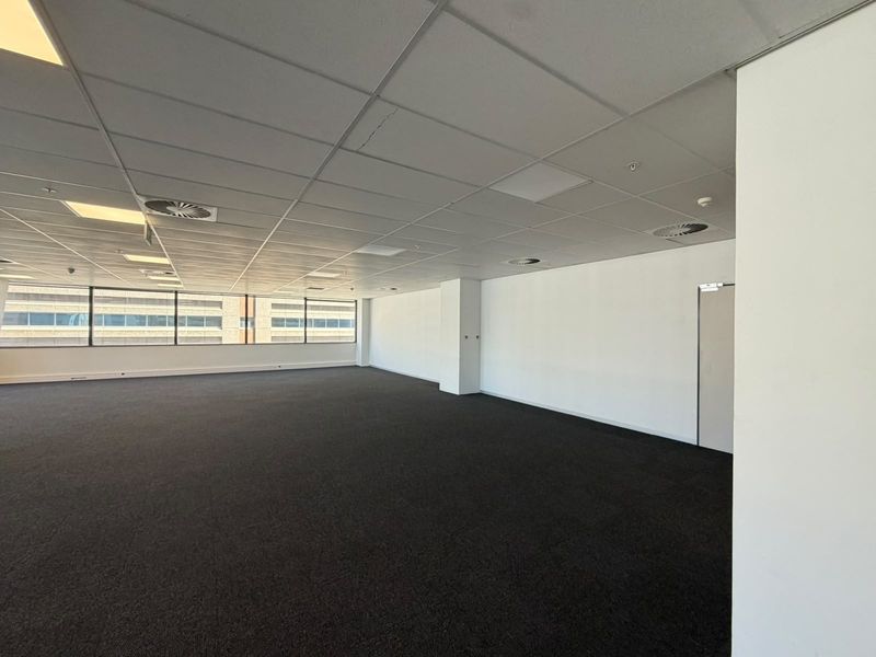 A spacious, empty open-plan office area featuring a suspended ceiling, recessed lighting, air conditioning vents, and large windows providing natural light.
