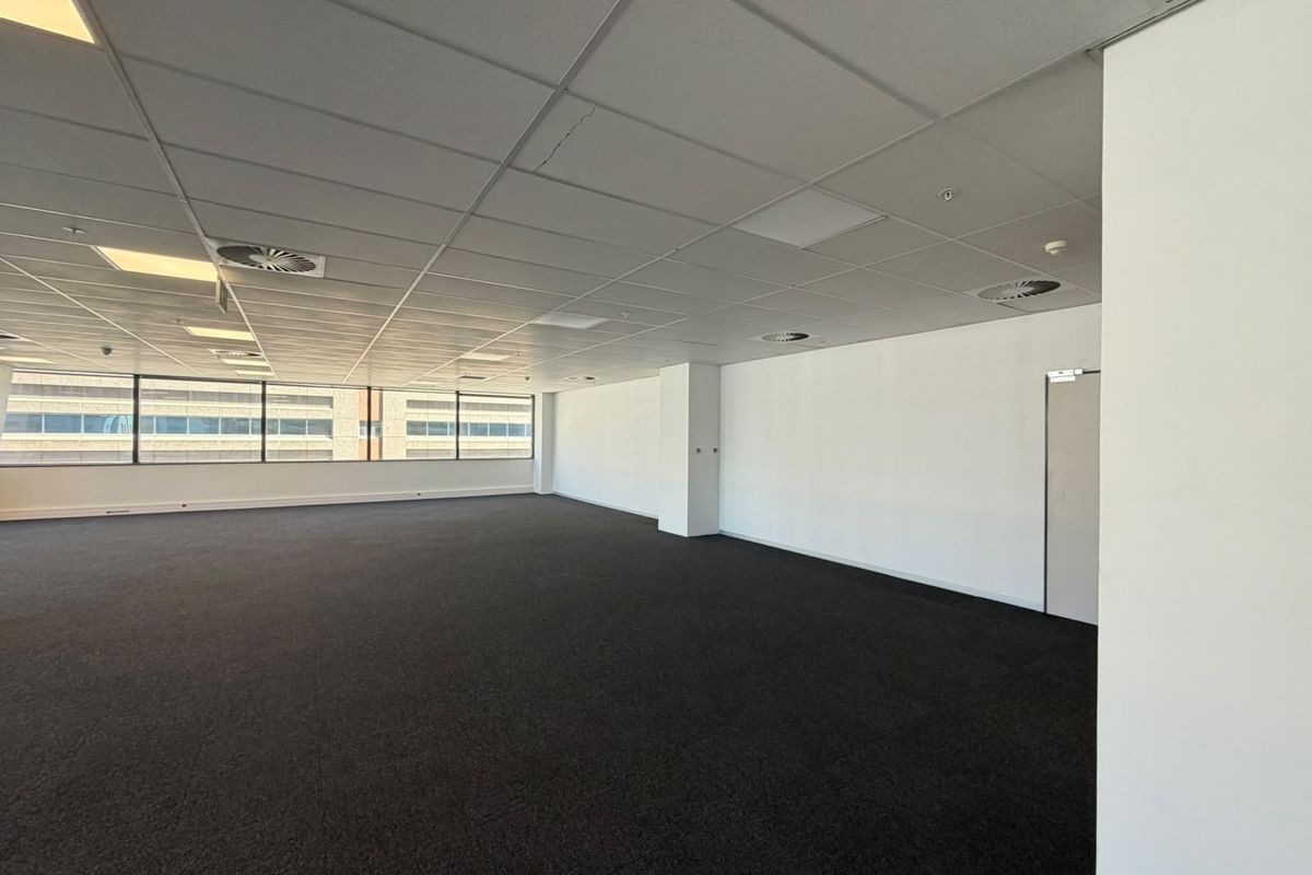 A spacious, empty open-plan office area featuring a suspended ceiling, recessed lighting, air conditioning vents, and large windows providing natural light.