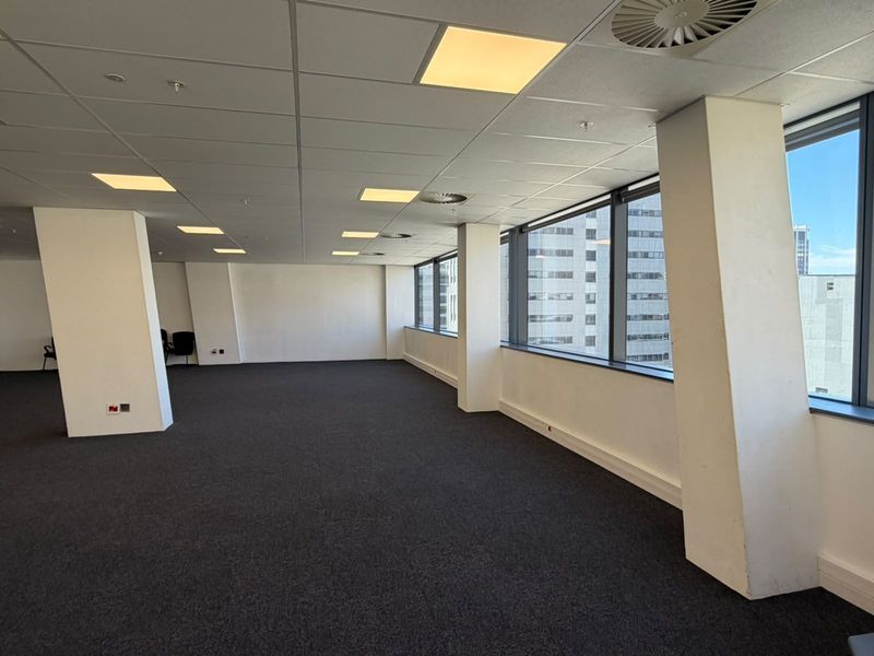 A spacious, open-plan office area featuring large windows, carpeted flooring, and a suspended ceiling with integrated lighting and air conditioning.